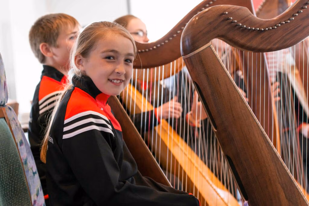A group of young people seated indoors playing harps, with one harp and a player who is smiling clearly visible in the foreground.