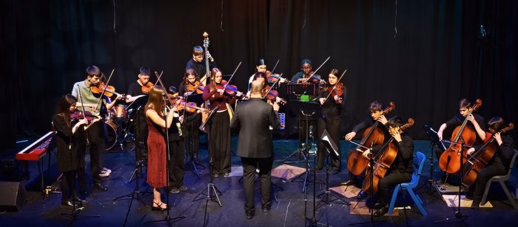 A group of young musicians with violins, violas, cellos and double basses performing on stage led by their conductor Davide Forti.