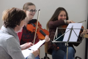 Participants involved in a music practice session, including two violin players and composer Elaine Agnew reviewing sheet music.