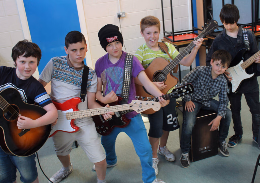 A group of young musicians posing with electric and acoustic guitars in a classroom music setting.