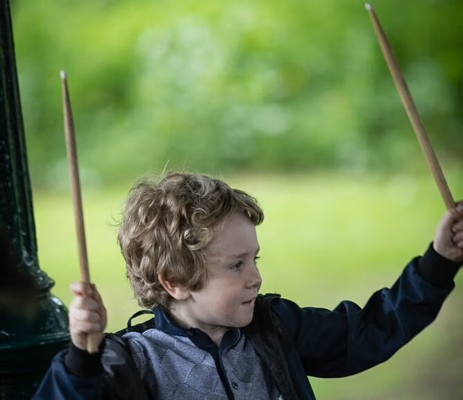 A student drummer holding drumsticks up high, practising rhythm skills on a drum kit at the band stand in St Helena’s park, Dundalk.