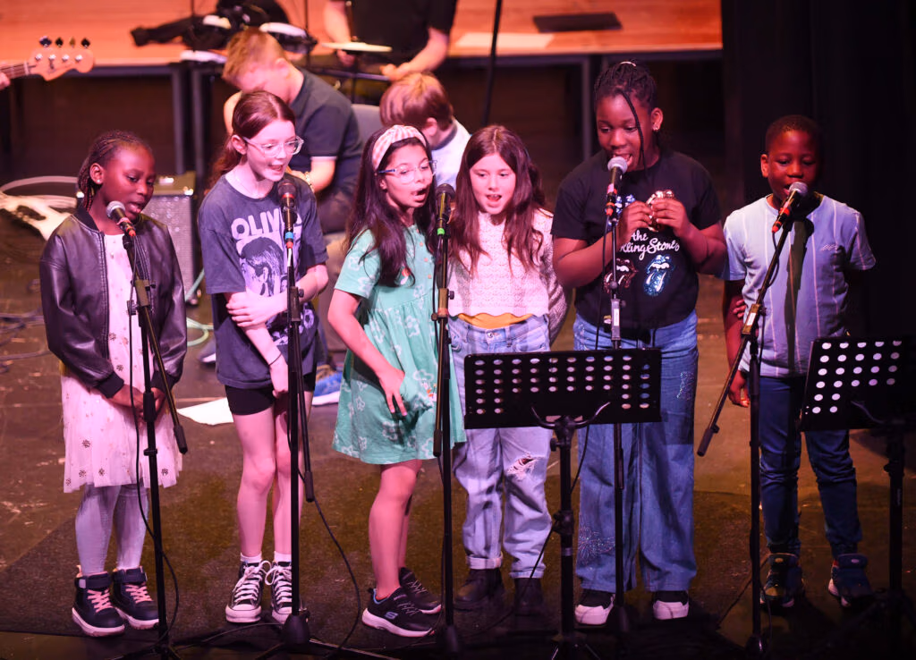 A group of six young people standing at microphones singing on stage with two guitarists and a drummer in the background.