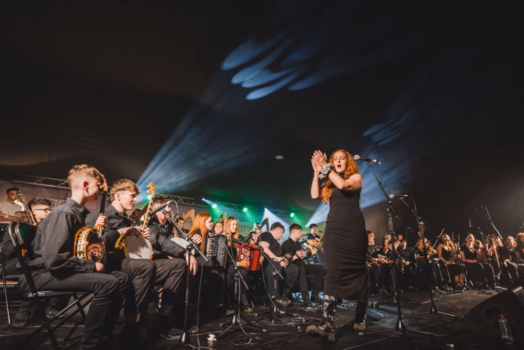 A large group of young musicians performing on stage at Fleadh Choeil na hÉireann playing a range of instruments including banjo, accordion, fiddle, concertina and pipes. They are being led by musician Lisa Canney who is leading from the front under concert lighting