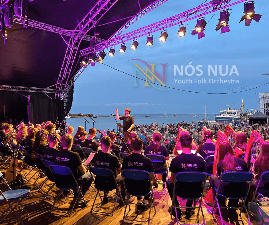 Nós Nua Youth Folk Orchestra performing in the early evening on an outdoor stage by the waterfront in Wexford during Fleadh Cheoil na nÉireann. They are led by a conductor under stage lighting and there is a large audience watching.