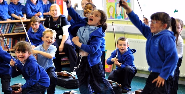 A group of primary school children in school uniform smiling and holding violin bows whilst actively participating in a classroom music session.
