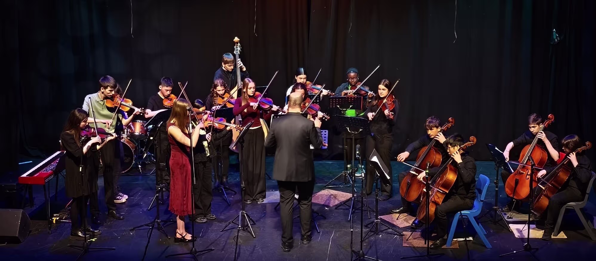 A group of young musicians with violins, violas, cellos and double basses performing on stage led by their conductor Davide Forti.