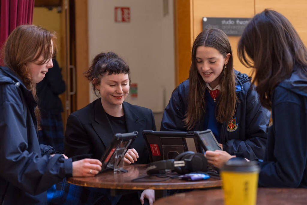 3 students with tablets are seated around a table discussing their musical tracks with guest musician Æ MAK aka Aoife McCann.
