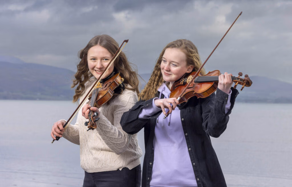Two young musicians playing violins together at Blackrock sea-front with Cooley hills in the background.