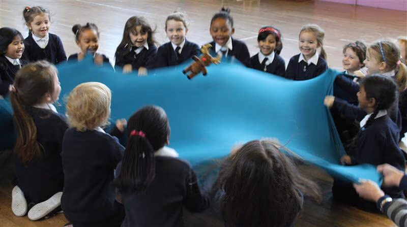 A group of young children gathered in a circle indoors part taking in a interactive early years music session using a shared fabric for movement and play.