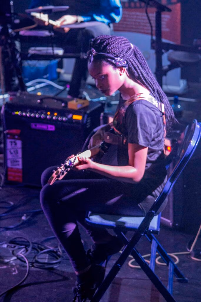 A young female guitarist performing on stage as part of a band, with drum kit and amplifiers in the background.”